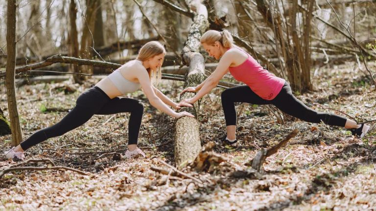 Side view of young female athletes in activewear stretching legs together using fallen tree during fitness workout in spring forest adhering to healthy lifestyle